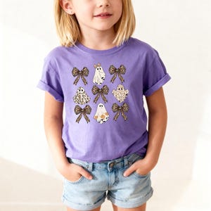 A young girl wearing a purple t-shirt with a graphic design of ghostly figures and leopard print bows.