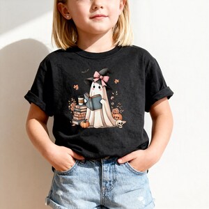 A young girl wearing a black t-shirt with a graphic of a ghost reading a book and surrounded by Halloween-themed decorations.