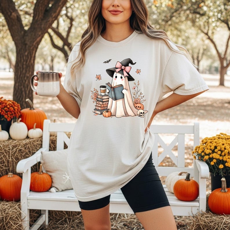 A woman wearing a white t-shirt with a graphic of a ghost reading a book, standing in front of a white bench with pumpkins and flowers.