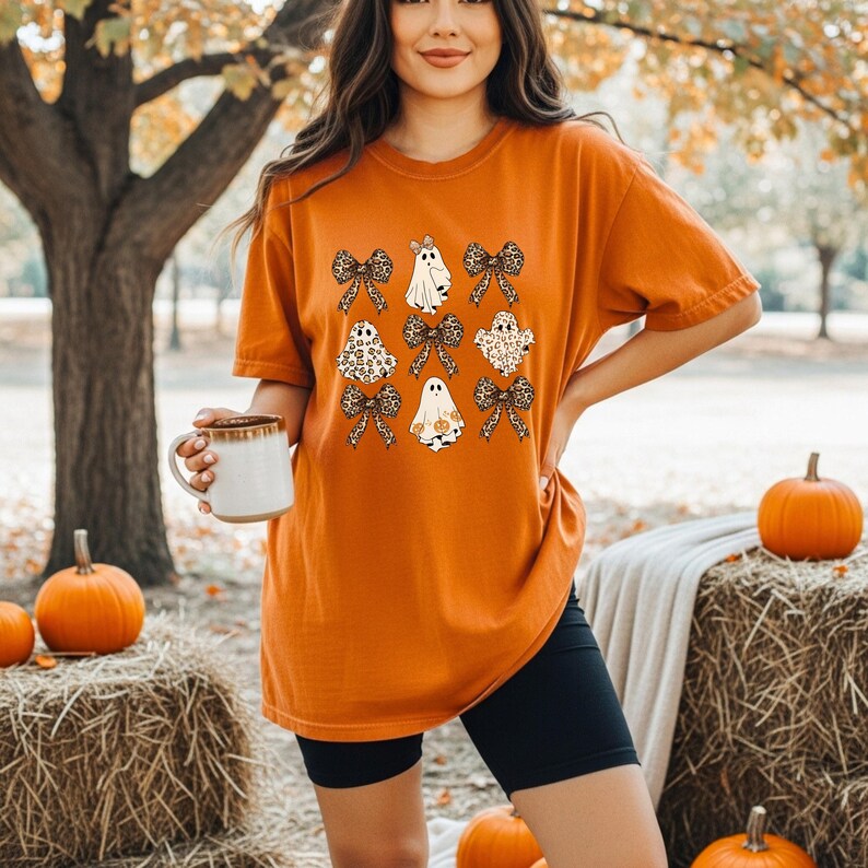 A woman wearing an orange t-shirt with a graphic of ghostly figures and bows stands in a park surrounded by hay bales and pumpkins, holding a mug.