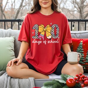 A woman wearing a red t-shirt with a graphic of a bow and the text "100 days of school" is sitting on a couch with a mug in her hand.