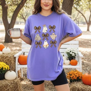 A woman wearing a purple t-shirt with a graphic of ghostly figures and bows stands in front of a white bench and pumpkins, holding a mug.