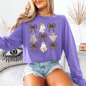A woman wearing a purple long-sleeved shirt with a graphic of ghostly figures and leopard-print bows, paired with denim shorts.