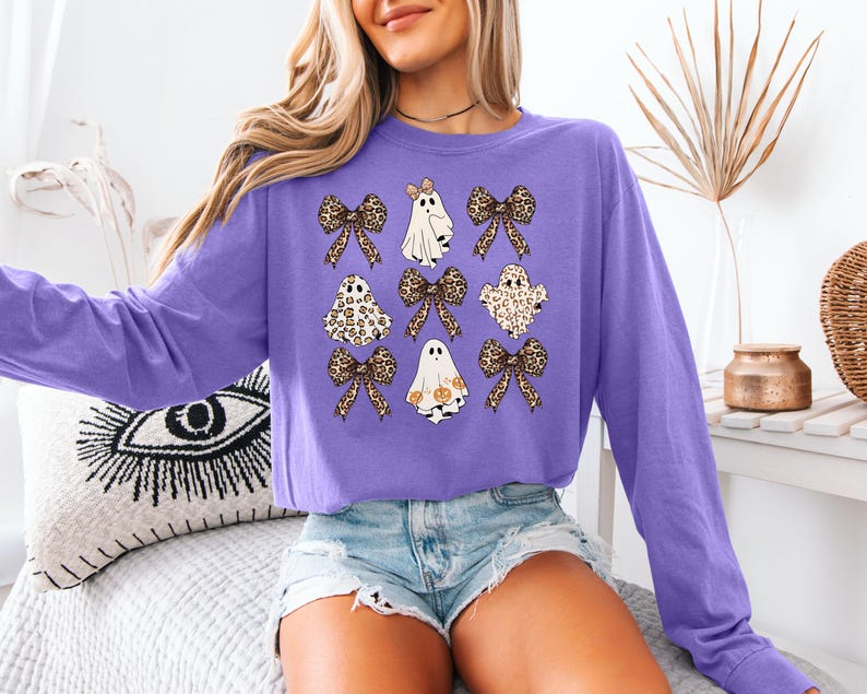 A woman wearing a purple long-sleeved shirt with a graphic of ghostly figures and leopard-print bows, paired with denim shorts.