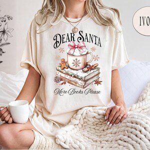 A woman wearing a white t-shirt with a graphic design of a mug and books, sitting on a bed with a white blanket.