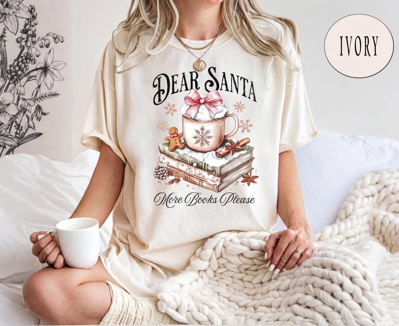 A woman wearing a white t-shirt with a graphic design of a mug and books, sitting on a bed with a white blanket.