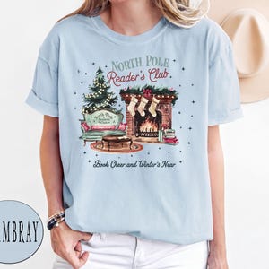 A woman wearing a light blue t-shirt with a Christmas-themed graphic design, including a fireplace, Christmas tree, and other holiday decorations.