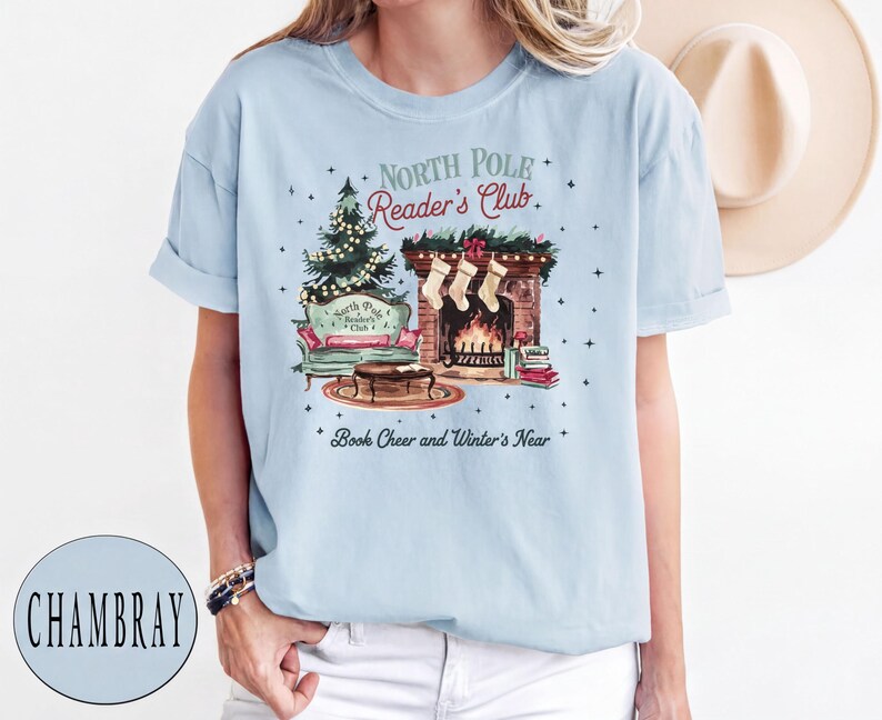 A woman wearing a light blue t-shirt with a Christmas-themed graphic design, including a fireplace, Christmas tree, and other holiday decorations.