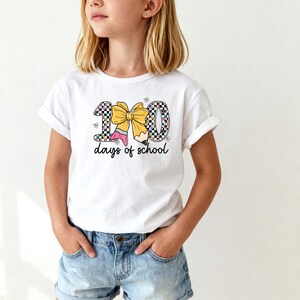 A young girl wearing a white t-shirt with a graphic design of a yellow bow and the text "100 days of school" printed on it.
