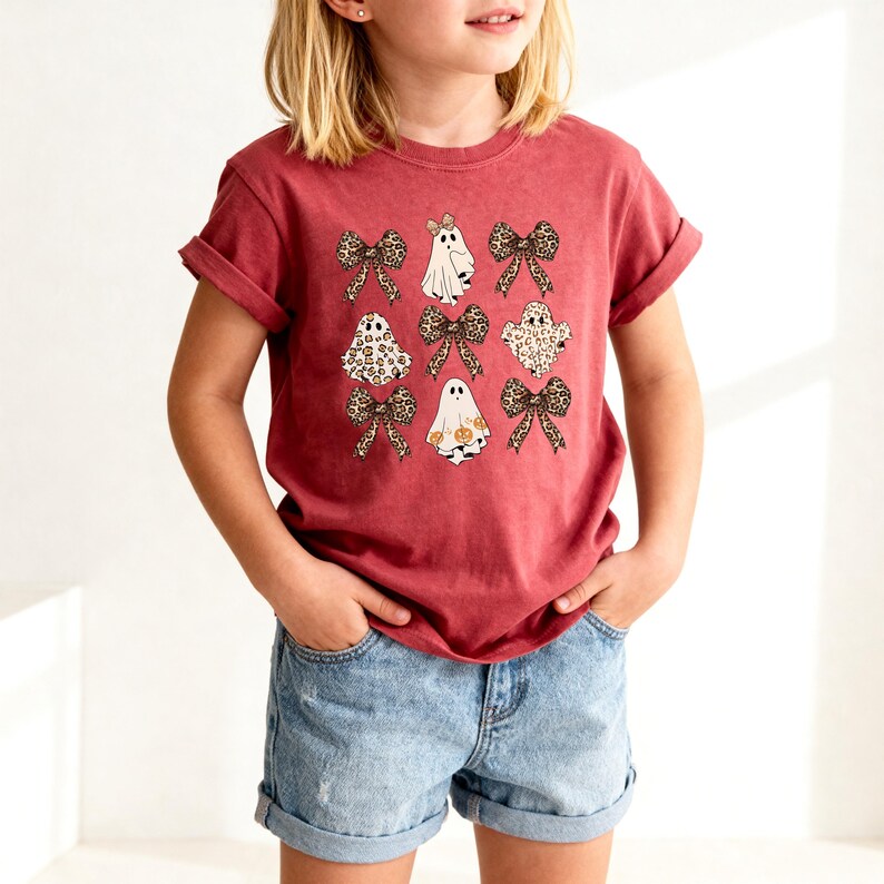 A young girl wearing a red t-shirt with a graphic design of ghostly figures and leopard-print bows.