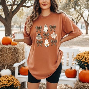 A woman wearing a t-shirt with a graphic of ghostly figures and bows stands in a park surrounded by pumpkins and hay bales.