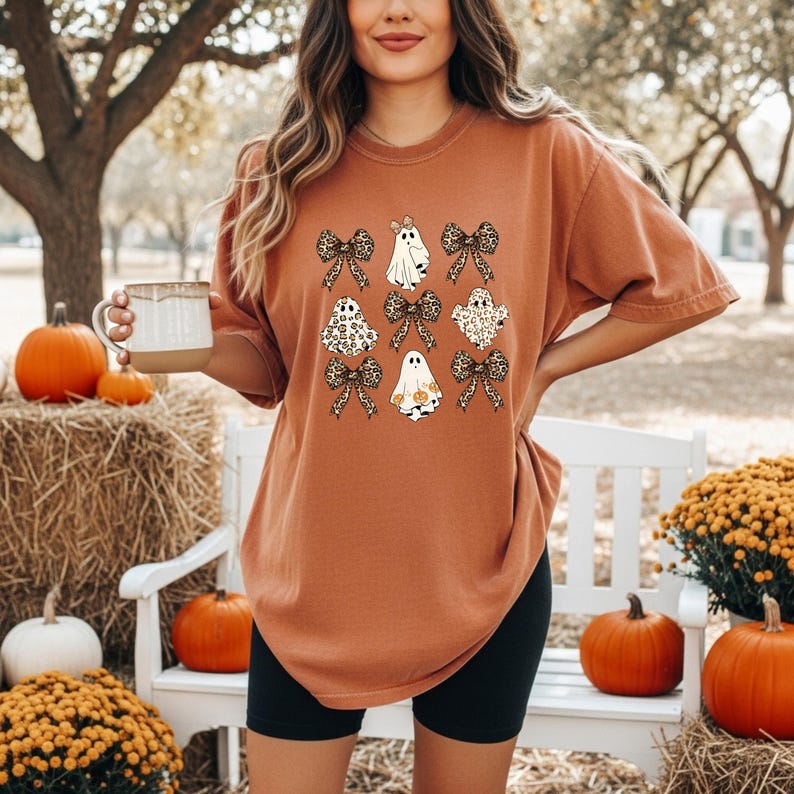 A woman wearing a t-shirt with a graphic of ghostly figures and bows stands in a park surrounded by pumpkins and hay bales.