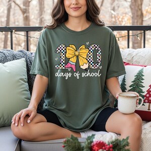 A woman sitting on a couch with a green t-shirt that says "100 days of school" and a bow on it. She is holding a mug in her hand.