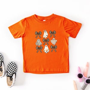 a bright orange t-shirt with a graphic of ghostly figures and bows, along with a pair of black and white checkered shoes and some children's art supplies, such as crayons and markers, arranged on a white background.