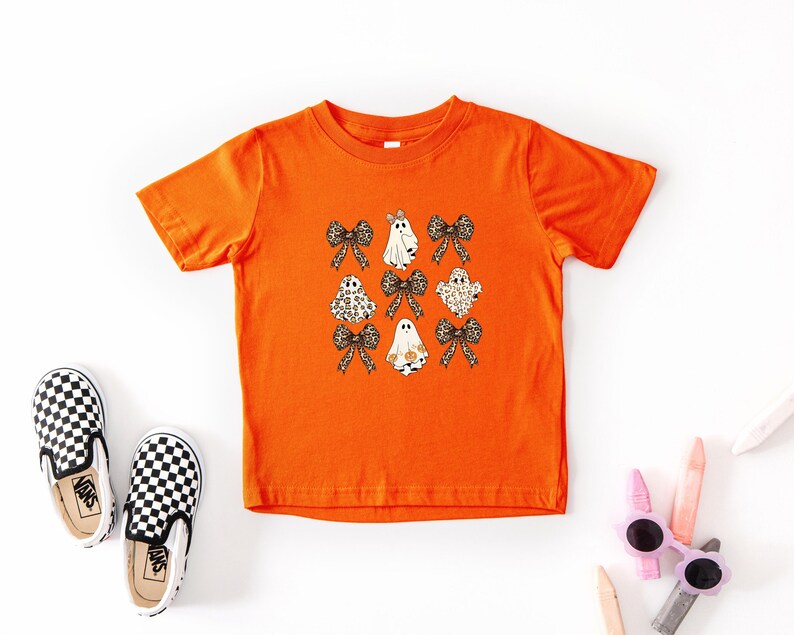 a bright orange t-shirt with a graphic of ghostly figures and bows, along with a pair of black and white checkered shoes and some children's art supplies, such as crayons and markers, arranged on a white background.