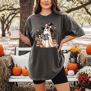 A woman wearing a t-shirt with a graphic of a ghost reading a book, holding a mug, and surrounded by pumpkins and flowers in an outdoor setting.