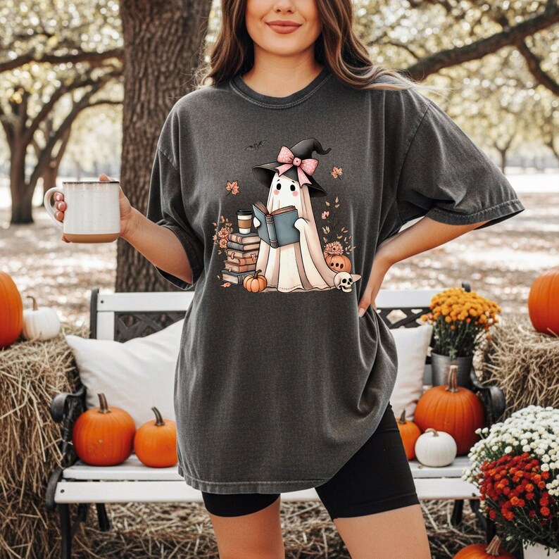 A woman wearing a t-shirt with a graphic of a ghost reading a book, holding a mug, and surrounded by pumpkins and flowers in an outdoor setting.