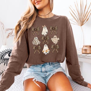A woman wearing a brown sweatshirt with a graphic of ghostly figures and leopard print bows, paired with denim shorts, sits on a bed with a white pillow.
