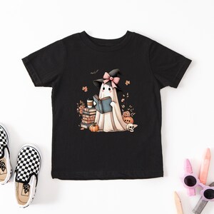 A black t-shirt with a graphic of a ghost reading a book, surrounded by Halloween-themed elements such as pumpkins, bats, and a witch's hat.