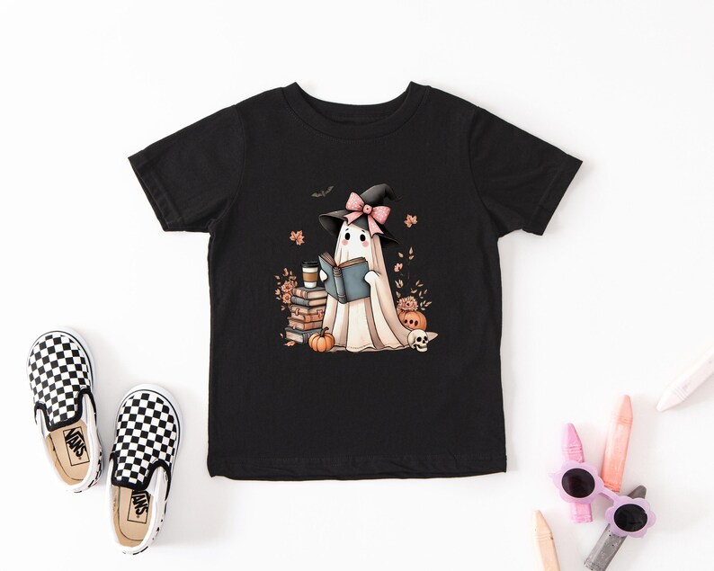 A black t-shirt with a graphic of a ghost reading a book, surrounded by Halloween-themed elements such as pumpkins, bats, and a witch's hat.