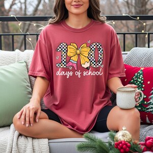 A woman sitting on a couch with a mug in her hand, wearing a red t-shirt with a graphic design and text.