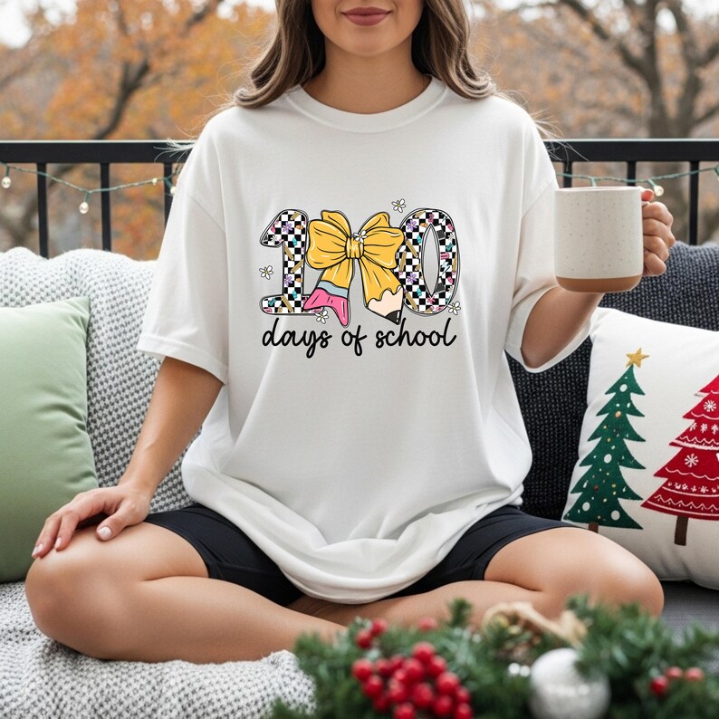 A woman sitting on a couch, wearing a white t-shirt with a graphic design of the number 100 and the text "100 days of school". She is holding a coffee mug in her hand.