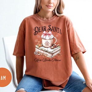 A woman wearing a t-shirt with a Christmas-themed design, including a mug, books, and snowflakes.