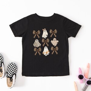 a black t-shirt with a Halloween-themed design, including ghost and bow motifs, along with a pair of black and white checkered shoes and a pair of pink and white sunglasses.