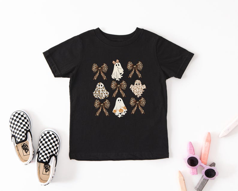 a black t-shirt with a Halloween-themed design, including ghost and bow motifs, along with a pair of black and white checkered shoes and a pair of pink and white sunglasses.