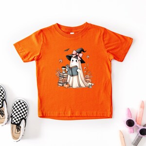 a bright orange t-shirt with a cartoon ghost wearing a witch's hat and holding a book, surrounded by Halloween-themed elements such as pumpkins, bats, and a skeleton.