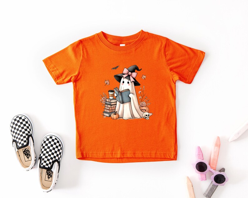 a bright orange t-shirt with a cartoon ghost wearing a witch's hat and holding a book, surrounded by Halloween-themed elements such as pumpkins, bats, and a skeleton.