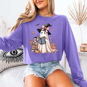 A woman wearing a purple long-sleeved shirt with a graphic of a ghost reading a book, surrounded by Halloween-themed decorations.