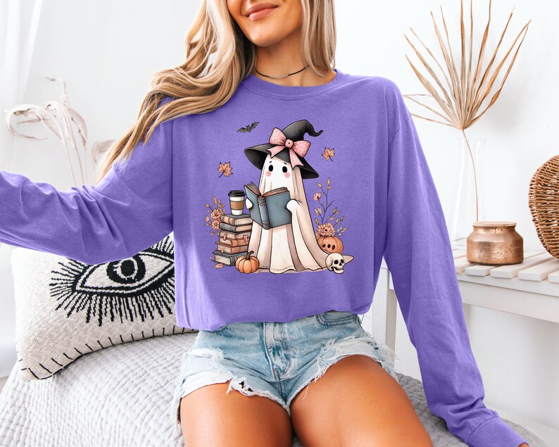 A woman wearing a purple long-sleeved shirt with a graphic of a ghost reading a book, surrounded by Halloween-themed decorations.