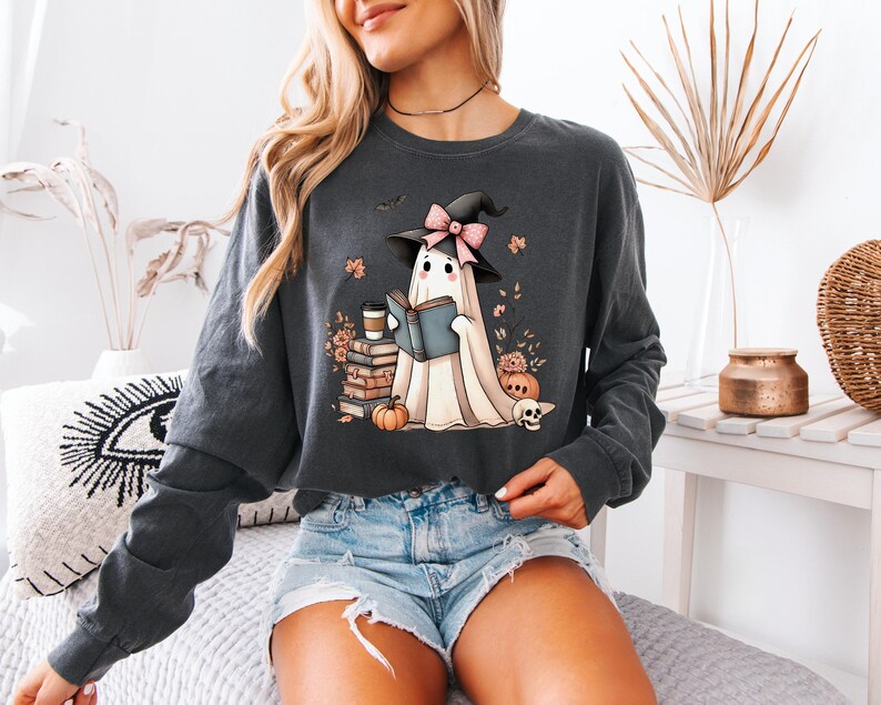 A woman wearing a gray sweatshirt with a graphic of a ghost reading a book, surrounded by Halloween-themed elements such as pumpkins, bats, and a witch's hat.