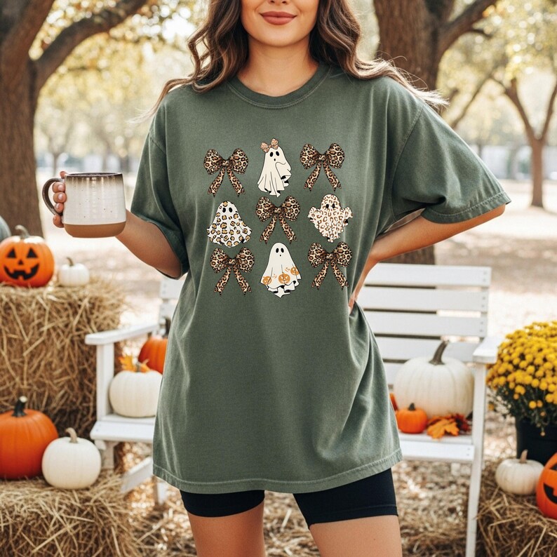 A woman wearing a green t-shirt with a graphic of ghostly figures and leopard-print bows, holding a mug, and surrounded by Halloween decorations such as pumpkins and hay bales.