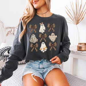 A woman wearing a gray sweatshirt with a graphic design of ghostly figures and leopard-print bows, paired with denim shorts.