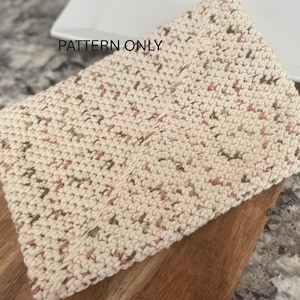 May include: A close-up of a crocheted dishcloth with a beige background and a speckled pattern of brown, green, and pink yarn. The dishcloth is lying on a wooden surface. "PATTERN ONLY" is written in black text on a white background in the upper left corner of the image.