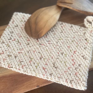 May include: A crocheted pot holder with a speckled pattern in beige, pink, and green. The pot holder is lying on a wooden surface.