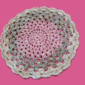 May include: A handmade, circular crochet doily with a scalloped edge. The doily features concentric circles of color, transitioning from a soft pink center to gray and beige tones on the outer edge. The background is a solid pink.