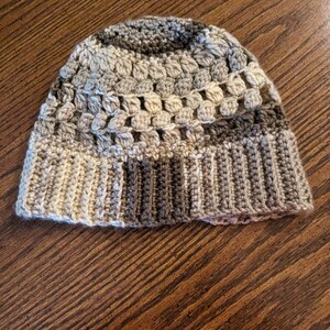 May include: A handmade crochet beanie in neutral tones of beige, brown, and gray. The hat features a ribbed brim and a textured stitch pattern on the crown. The color variations create a striped effect, offering a cozy and stylish accessory.