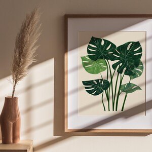 May include: A framed print of a watercolor illustration of a green monstera plant on a cream background.
