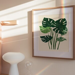 May include: A framed print of a botanical illustration featuring a cluster of green monstera leaves on a cream background.
