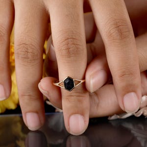 May include: A gold ring with a black hexagonal stone set in the center. The ring is on a finger.