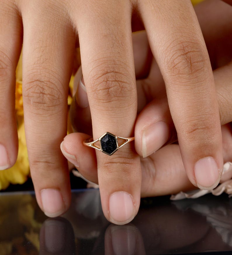 May include: A gold ring with a black hexagonal stone set in the center. The ring is on a finger.