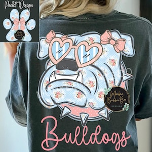May include: A charcoal gray t-shirt featuring a large graphic on the back of a bulldog wearing heart-shaped glasses and a bow. The bulldog has a light blue polka dot and floral pattern. The word Bulldogs is printed in pink below the graphic.
