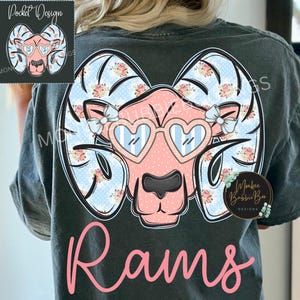 May include: Dark gray t-shirt featuring a pink ram design wearing heart-shaped sunglasses. The ram's horns are decorated with floral patterns and blue polka dots. The word "Rams" is written in pink script below the design.