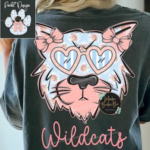 May include: A charcoal gray t-shirt featuring a large graphic of a cat face wearing heart-shaped glasses and bows on its ears. The cat has a blue polka dot and floral pattern with pink accents. The word Wildcats is printed in pink script below.