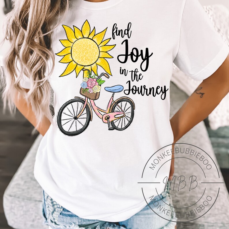 May include: White t-shirt with a graphic of a yellow sunflower, a pink bicycle with a basket of flowers, and the text "find joy in the journey".
