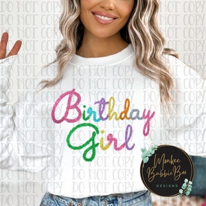 May include: A white sweatshirt with the text "Birthday Girl" in rainbow glitter lettering. The text is in a cursive font. The sweatshirt is being worn by a person.