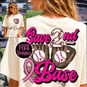 May include: A cream-colored t-shirt featuring a graphic print on the back with the text "Save 2nd Base" in bold pink letters. The design includes two brown baseball gloves, two baseballs with pink stitching, and a pink awareness ribbon.
